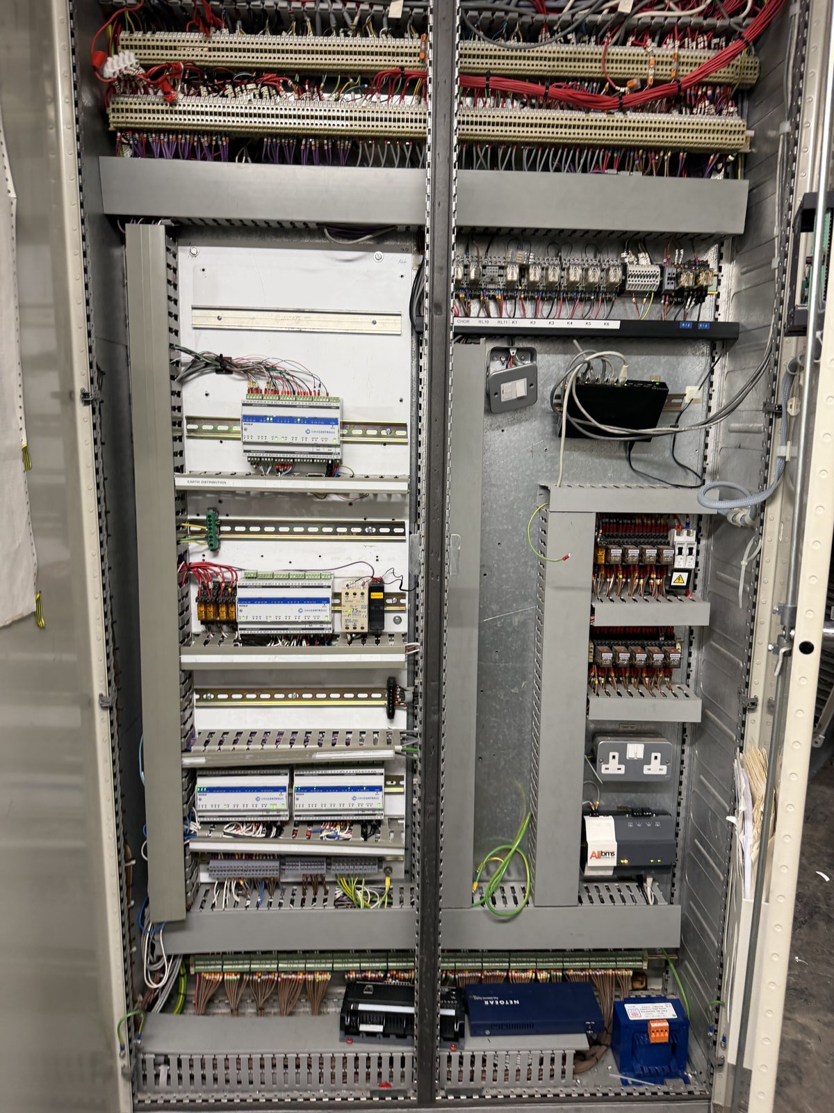 Interior of a building management system control panel with DIN-rail controllers, relays, terminals and a network switch, serving fan and plant control